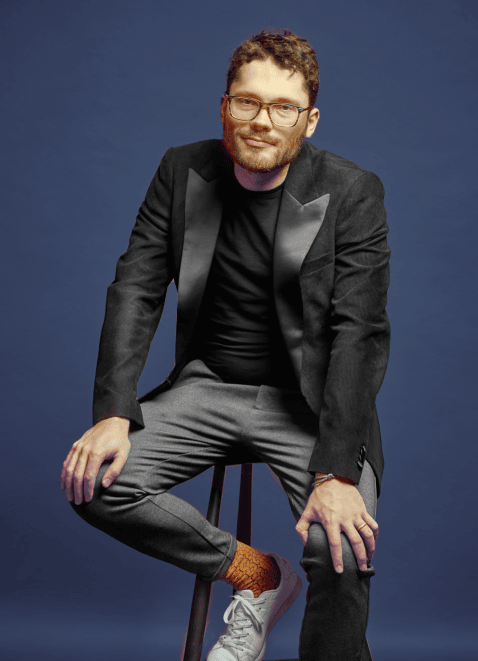 Adrian Goldner in schwarzem Sakko auf einem Barhocker vor blauem Hintergrund