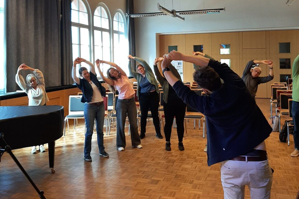 Workshop-Teilnehmende im Saal beugen sich mit über dem Kopf verschränkten Armen zur Seite